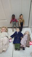 Five collectible dolls placed on floor against a door including large dolls and figures with clothing.