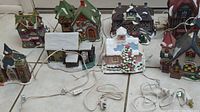 Eight Christmas village houses arranged on tiled floor with cords and plugs visible for lighting.