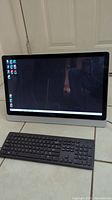 Front view of Dell Inspiron all-in-one computer powered on with Windows desktop on screen, wireless keyboard visible in front.
