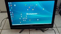 Full front view of Lenovo all-in-one desktop powered on showing desktop screen and Lenovo logo below screen.