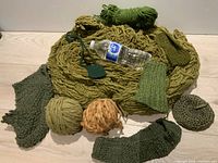 View of various knitting projects and yarn skeins laid out with water bottle for size reference; includes multiple green and beige yarn skeins and knitted pieces of various shapes and sizes