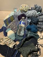 Top view of multiple knitting projects and yarn skeins showing variety in colors and materials with a water bottle for scale.