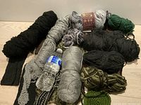 View of assorted yarn skeins and knitting projects showing black, gray, green yarns, and mixed color skeins laid out on surface with a water bottle for scale