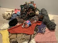 Multiple knitting projects and skeins of yarn piled together, showing various colors and stitches, including orange triangular piece, pink piece, grey skein and other smaller knitted portions.