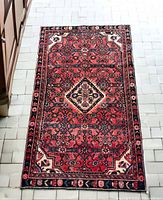 Top-down view of red Persian Hamadan rug on tiled floor showing central diamond medallion and border pattern