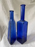 Photo shows two cobalt blue large glass bottles, one with square base taller than the other with a triangular base.