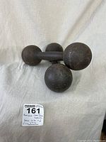 Two antique cast iron dumbbell-shaped weights with spherical ends and rusted surface, displayed on a neutral fabric.