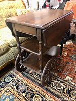 Side view of antique wooden tea trolley showing drop leaves, drawer, and large wooden wheels.