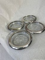 Four circular coasters with silver-tone stamped brass rims and embossed glass centers arranged with some overlapping and lay flat on beige fabric.