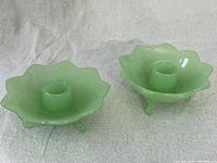Two light green jadeite glass candle holders with floral pattern and three legs, shown side by side on a light fabric background.