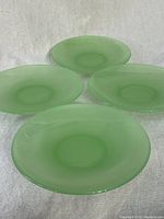 Top-down view showing all four jadeite glass tea saucers arranged on a white fabric surface, highlighting their pale green translucent color and circular shape.