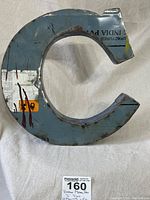 Front view of a large metal letter C art piece with visible welded edges and rust spots on faded paint.