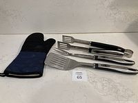 Photo shows black and blue long fabric BBQ mitt alongside stainless steel BBQ utensils: spatula, fork, and tongs.