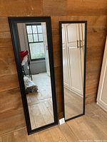 Photo showing both mirrors standing against a wooden wall on a hardwood floor. Left mirror with thick frame, right mirror with thin frame reflecting room details.