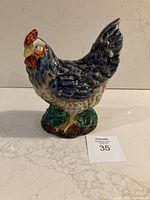 Front angled view of ceramic rooster figurine on white surface with auction lot number card visible.