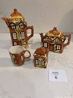 Four ceramic pieces designed as cottages: coffee pot, teapot, small container, and cup with detailed textures and paint.