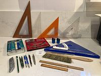 Full view of all drafting supplies including triangles, rulers, pens, compasses, ink bottles, and a drafting brush displayed on a flat surface.