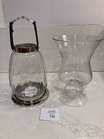 Two glass lanterns displayed together on a marble surface; one lantern has a silver metal base and blue faux leather handle, the other is a plain glass hurricane vase shaped lantern with pedestal base.