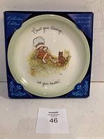 Front view of the Holly Hobbie collector's plate in original box. Plate shows a girl with a bonnet and a cat sitting on grass with the text "Count your blessings... not your troubles."
