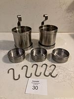 Two stainless steel hanging pots, three round tins with clear lids and magnets underneath, and five S-hooks lined up on a counter, all accessories included with the IKEA Grundtal shelf.