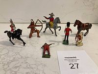 Photo showing all seven painted metal toy figurines: black horse soldier, archer, cowboy on gray horse, British guard, soldier lying down, brown horse, and small cannon.