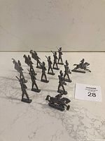 Overview photo showing 13 metal toy soldier figurines in various poses, some mounted on horseback, others with rifles, drums, and flags. Included is an auction lot card with number 28.