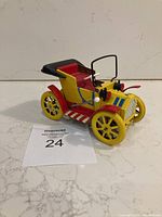 Front three-quarter view of yellow and red painted metal toy car resembling an early automobile, showing pull-back handle with white knob and yellow wheels.