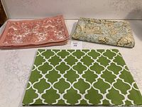 Eight orange color cotton placemats and four green placemats folded, alongside green tablecloth folded with botanical print