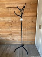 Full view of black forged metal coat tree standing against wooden wall with three-legged base and branch-like hooks.