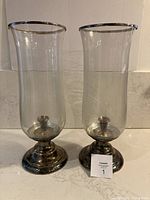 Two silver-plated taper hurricane lamps showing glass chimneys with silver rims and tarnished silver bases on a counter.
