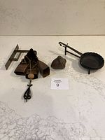 Three rustic metal accent pieces arranged on white marble surface: cowbell hanger with mounting bracket on left, acorn-shaped piece in center, small round scalloped dish on right.