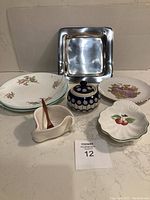 View of all seven items included in the lot arranged on a tabletop: two aqua edge floral plates, one gold edge plate, the scallop-shaped cherry dish, blue bowl with lid, the Hilborn pottery swirl dish with spoon, and square pewter plate.