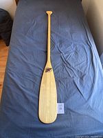 Full view of the paddle laid flat on a dark fabric surface, showing the front with the brand logo and natural light blonde wood finish.