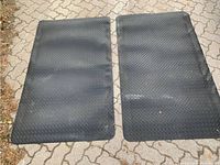 Pair of two large black rubber work station mats laid side by side, showing their checker plate patterned non-slip surfaces and rectangular shape.