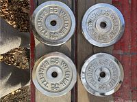Top view of four chromed barbell weight plates arranged in two rows on a wooden surface, showing their uniform size and marking.