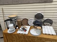 Setup showing most items on a gold tablecloth outside: coffee urn, drink mixer, canning pot, trays, dishes, skewers, knives and glass jars.