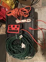 Overview photo showing the red power box, orange cable, white coiled extension cord, and two green extension cords all placed on a black plastic surface.