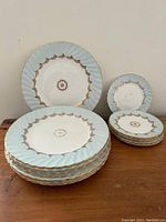 Stack of 8 large and 6 small bone china plates with pale blue rim and gold leaf detailing on wood surface.