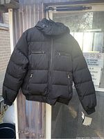 Front view of black puffy winter jacket with zipped pockets and hood hanging.