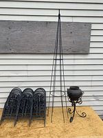 Photo showing 15 black steel lawn pickets stacked together, a 5-foot steel vine tower, and a black iron flowerpot holder on a stand with a round pot holder and scrollwork base.