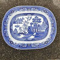 Large blue willow pattern serving platter photographed on a gravel ground for full view of design and shape.