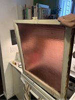Mauve/purple pebbled glass window held by hand, showing the textured glass and white painted wooden frame.