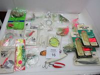 Photo of multiple fishing lures, teasers, hooks, and tackle components arranged on white background with auction card 16 in center. Items range in color and include some packaged and some loose in zip bags.