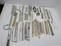 Wide view of over 30 metal watch bracelets laid out on a white surface showing a variety of silver and gold tone bands, different link styles and sizes.
