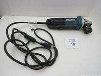 Makita electric angle grinder with attached power cord laid flat on white surface with auction lot card number 19.