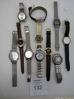 Full overview of 11 men's watches in various styles and bands laid flat on white surface.