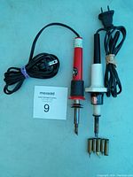 Two electric soldering irons with attached cords and additional tips on blue background.
