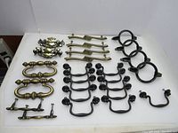 Photo showing full set of 25 drawer pullers arranged on white surface, displaying vintage brass-tone and black metal designs with original screws attached.