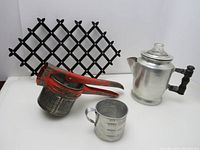 Shows all four items in the lot: foldable metal trivet, aluminum ricer with red handles, aluminum measuring cup, and coffee percolator with glass knob and wood handle.
