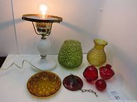 Working vintage milk glass hobnail lamp base with brass fittings and bulb illuminated. Several glass lamp shades and parts in green, yellow, red, and amber colors laid out beside it.